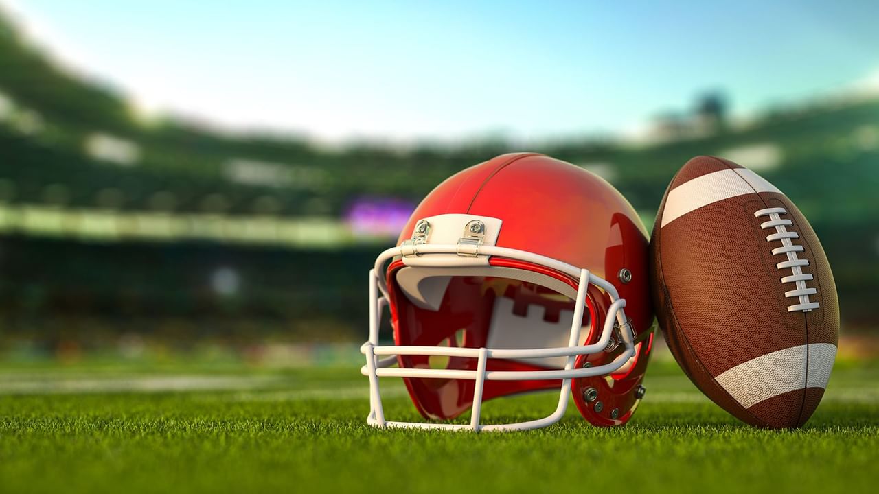 Red football helmet and brown football on green grass with blurred stadium in the background.