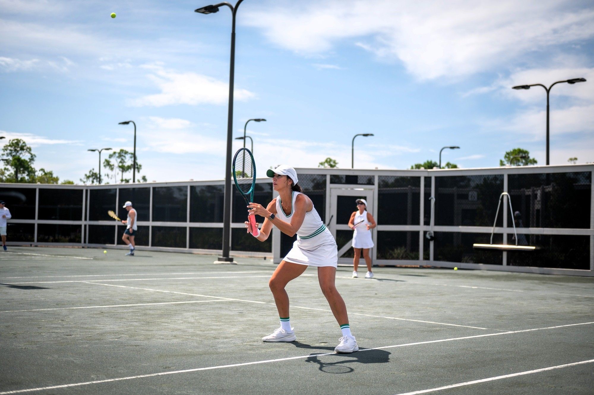 Woman in white tennis outfit holding racket preparing to hit ball on court with three other players.