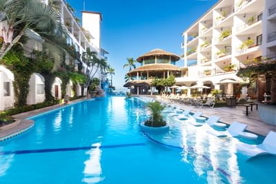 Bright blue pool with white lounge chairs surrounded by white hotel buildings and palm trees at Playa Los Arcos Puerto Vallarta