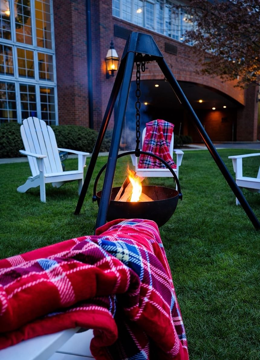 Outdoor tripod fire pit surrounded by white adirondack chairs with plaid blankets on lawn at Avon Old Farms Hotel
