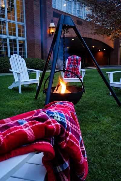 Outdoor tripod fire pit surrounded by white adirondack chairs with plaid blankets on lawn at Avon Old Farms Hotel