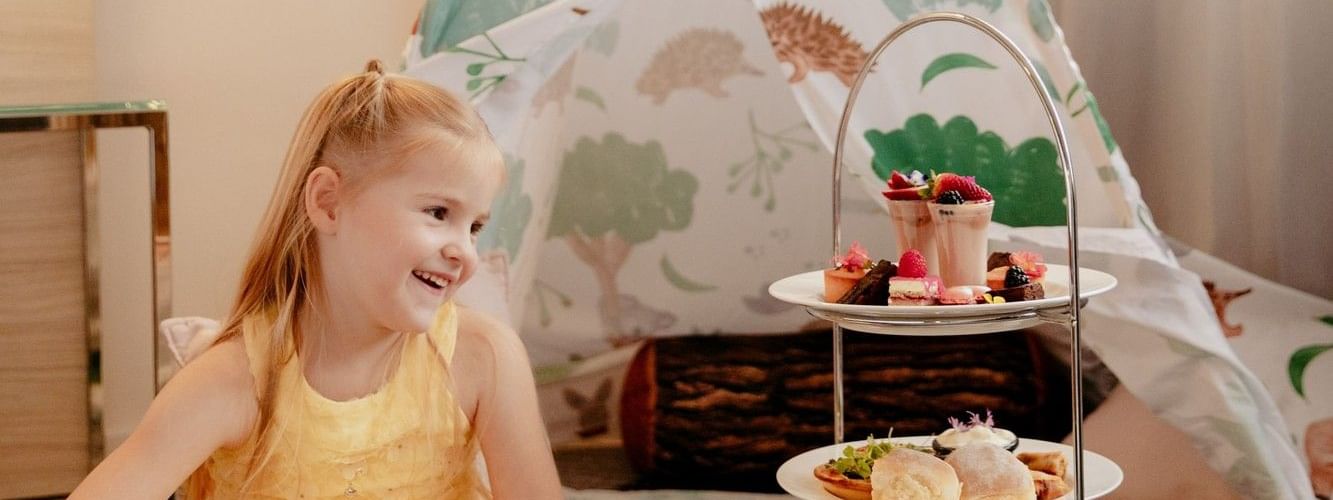 Smiling girl in a yellow dress at Novotel Sydney Parramatta, sitting near teepee and a tiered tray of desserts