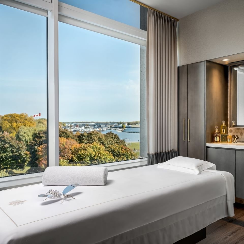 Spa treatment bed with rolled towel by a wardrobe, set under windows overlooking the marina view at Hotel X Toronto