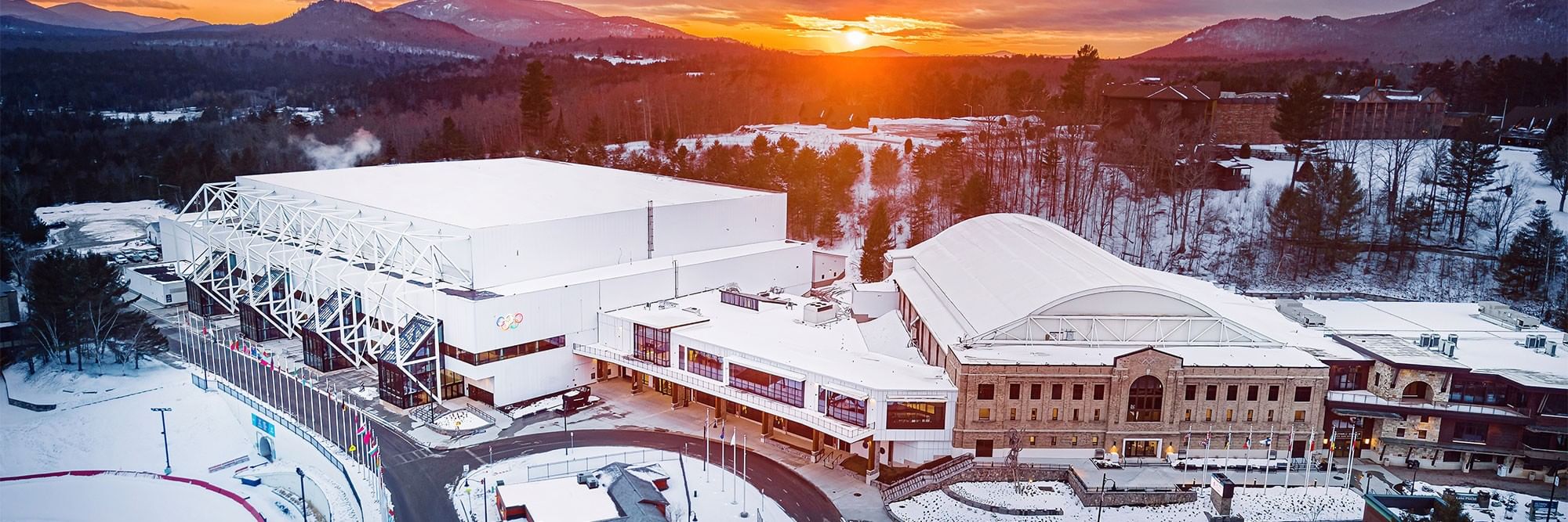 The image showcases a scenic view of a sports complex surrounded by snow-covered landscapes. A vibrant sunset casts warm hues over the buildings, highlighting the modern architecture of the arena and the historic structure nearby. The mountainous backdrop adds to the picturesque winter setting.