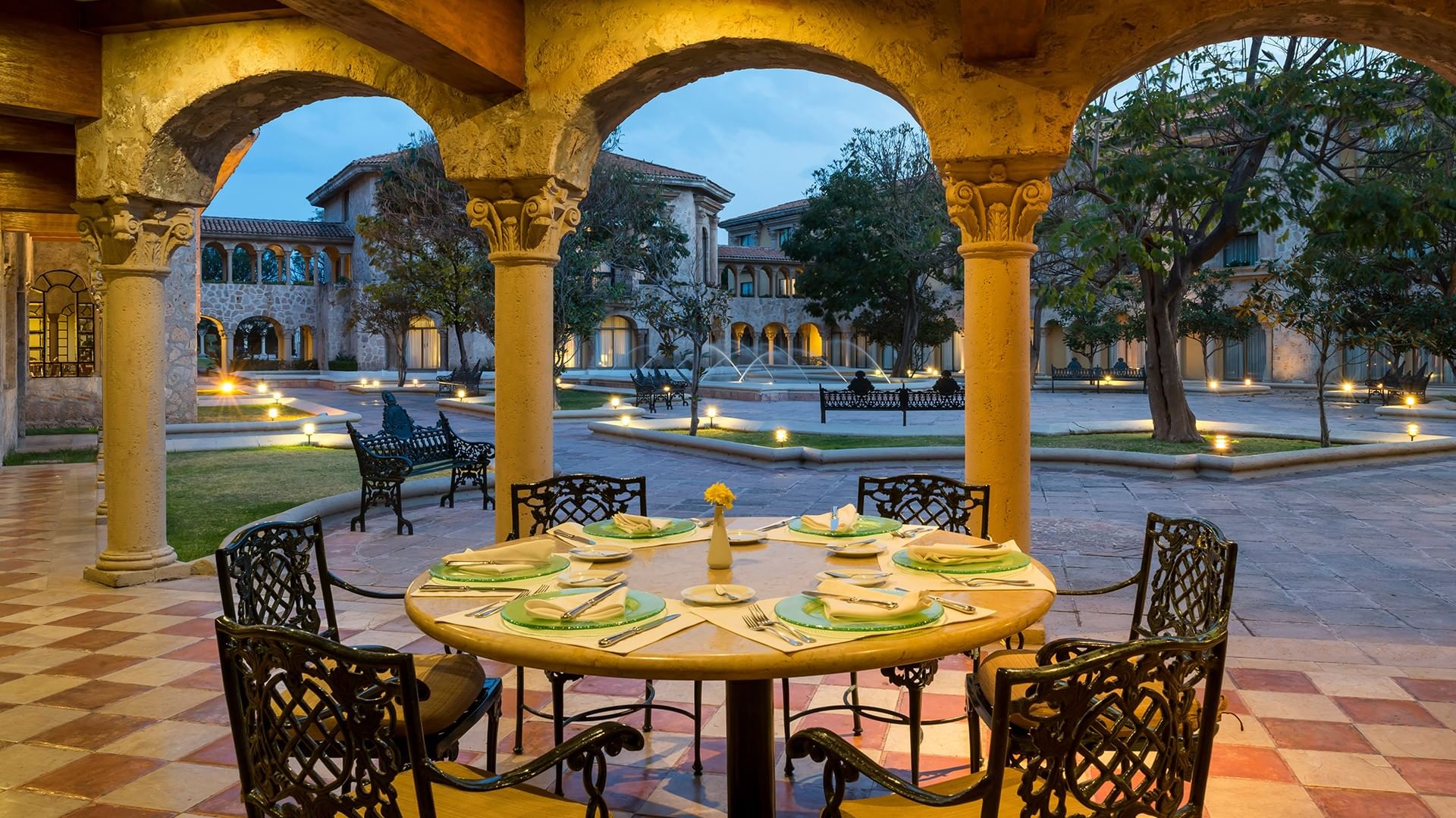 Mesa redonda con manteles verdes bajo arcos de piedra con vista a jardines iluminados en Quinta Real Aguascalientes