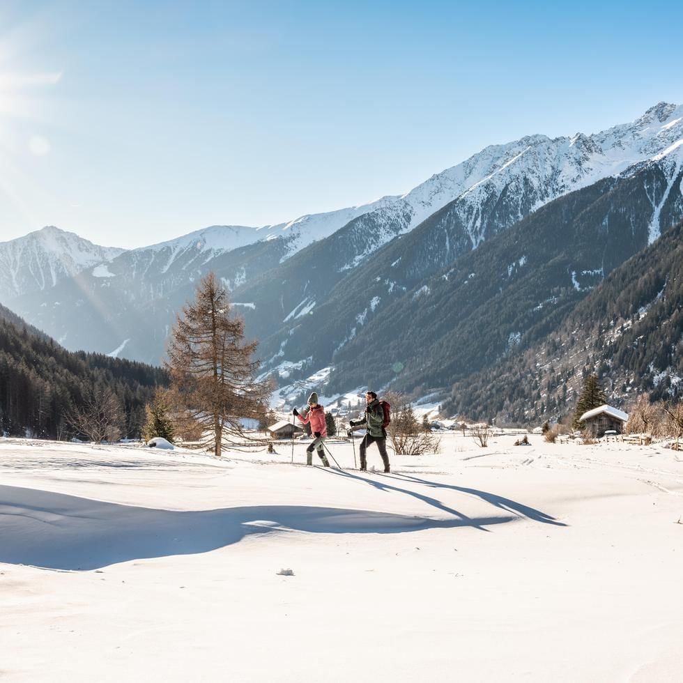 Zwei Wanderer mit Stöcken und Rucksäcken auf schneebedecktem Gelände vor verschneiten Bergen.