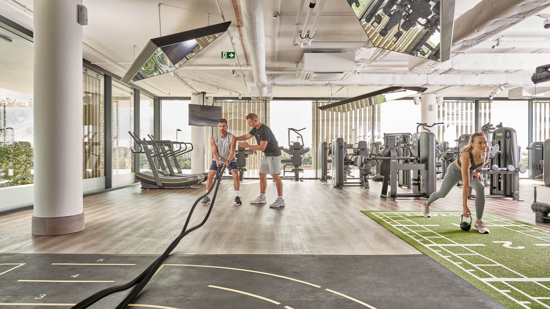 Drei Personen trainieren in einem modernen Fitnessstudio mit verschiedenen Geräten und einer hellen Atmosphäre.