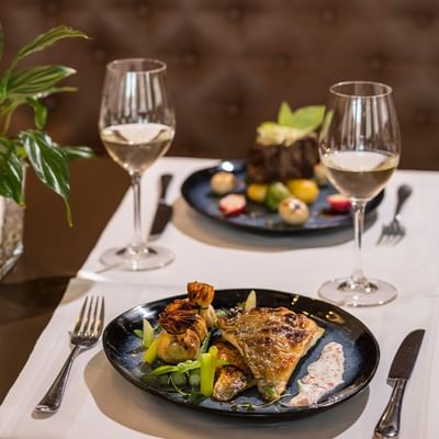 Zwei Teller mit Gerichten und Wein, Besteck und Pflanzen auf einem Tischtuch.