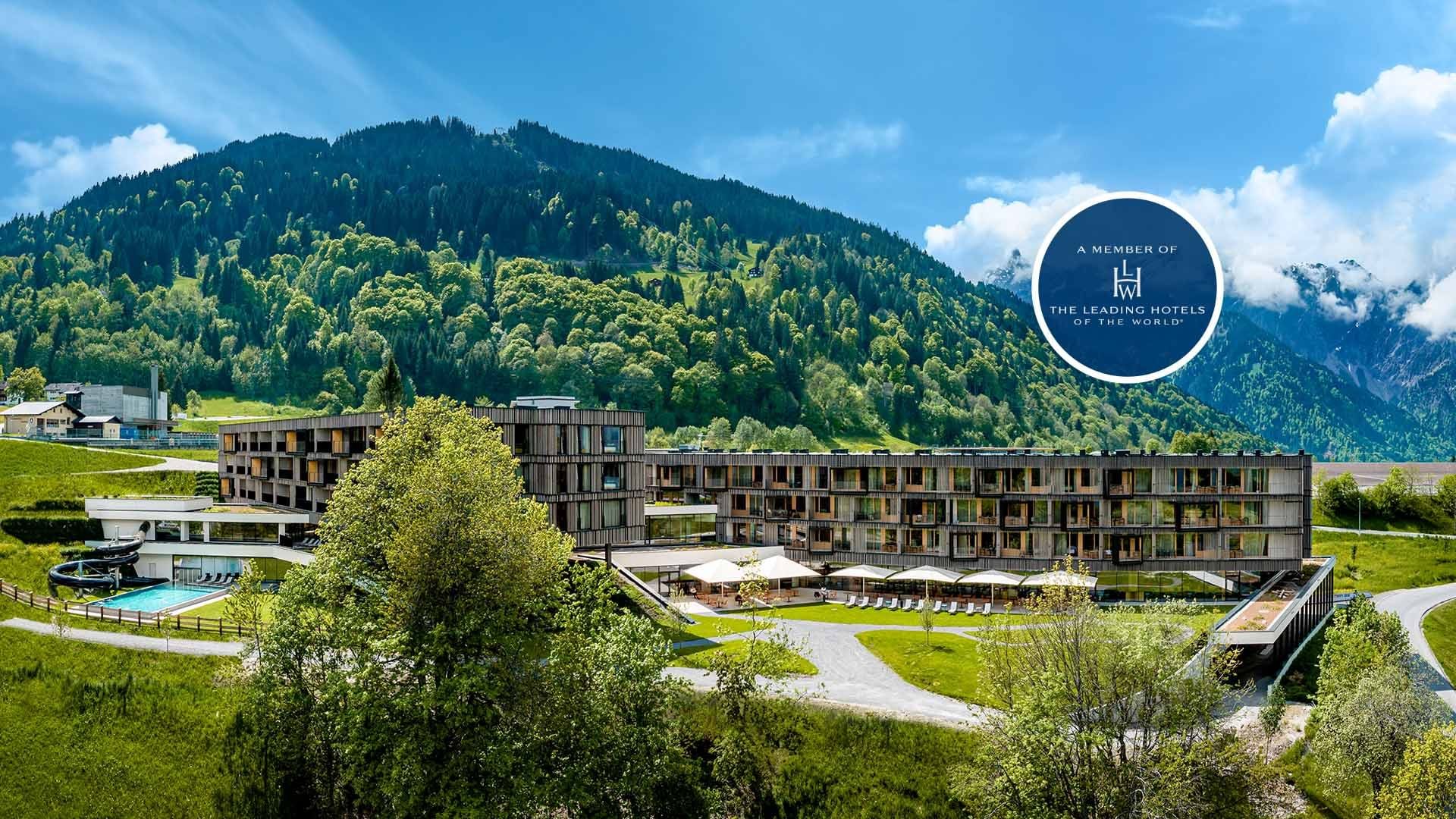 Modernes Hotelgebäude mit Pool, umgeben von grünen Wiesen und Bergen im Hintergrund.