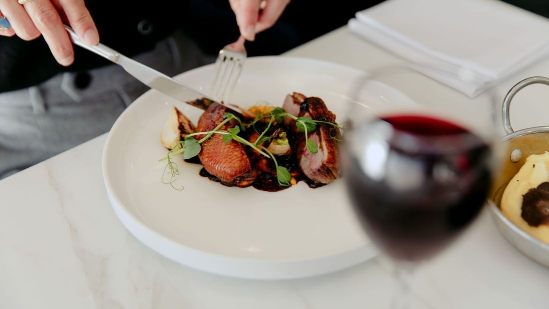 Person cutting meat on a plate with a glass of red wine and a bowl of food nearby.