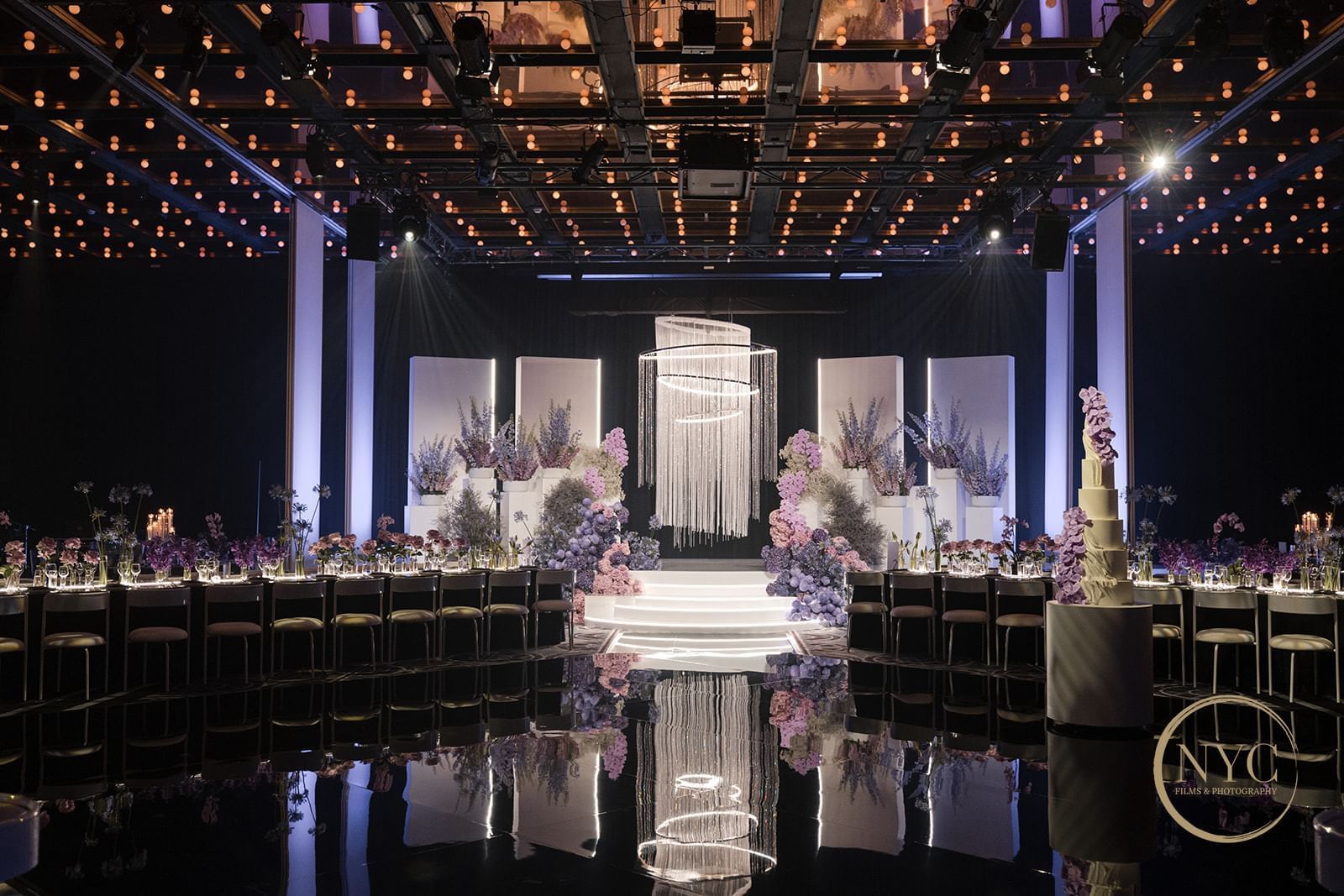 Decorated wedding reception hall with tables, flowers, a stage, and a cake, with a mirrored floor and spotlights.