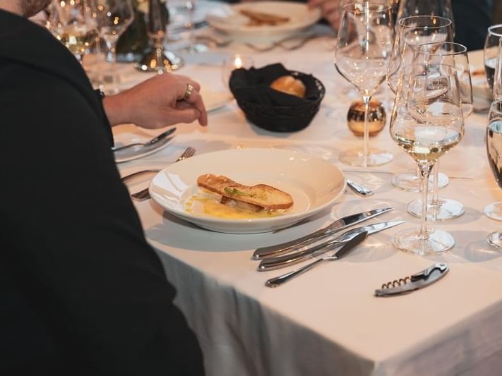 Person at a dining table with a plate of food, wine glasses, and cutlery.