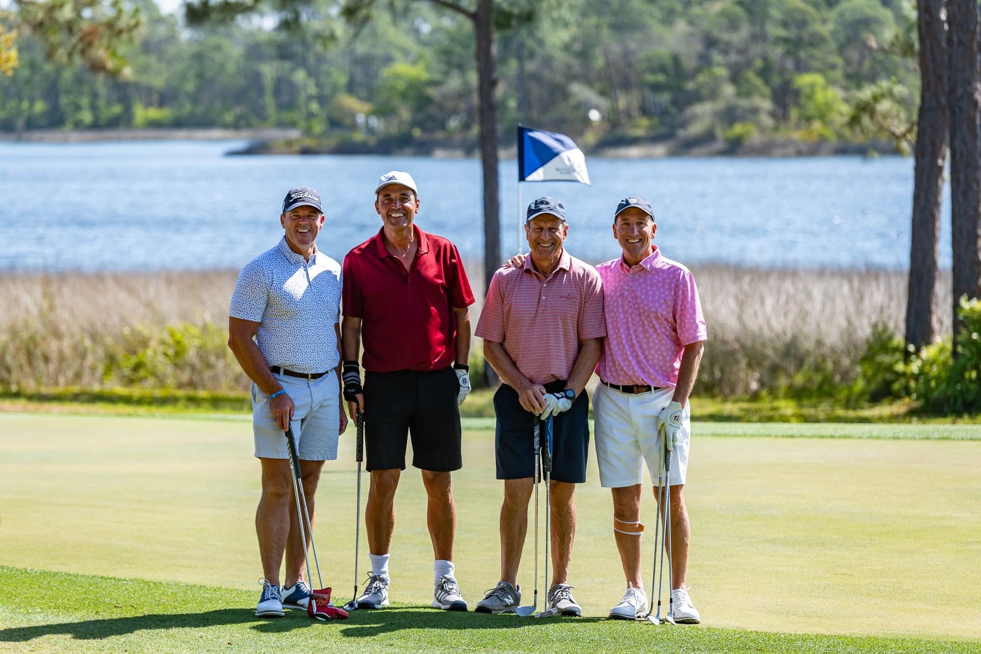 Four men in golf attire stand together on a golf course by a lake, smiling and holding golf clubs.