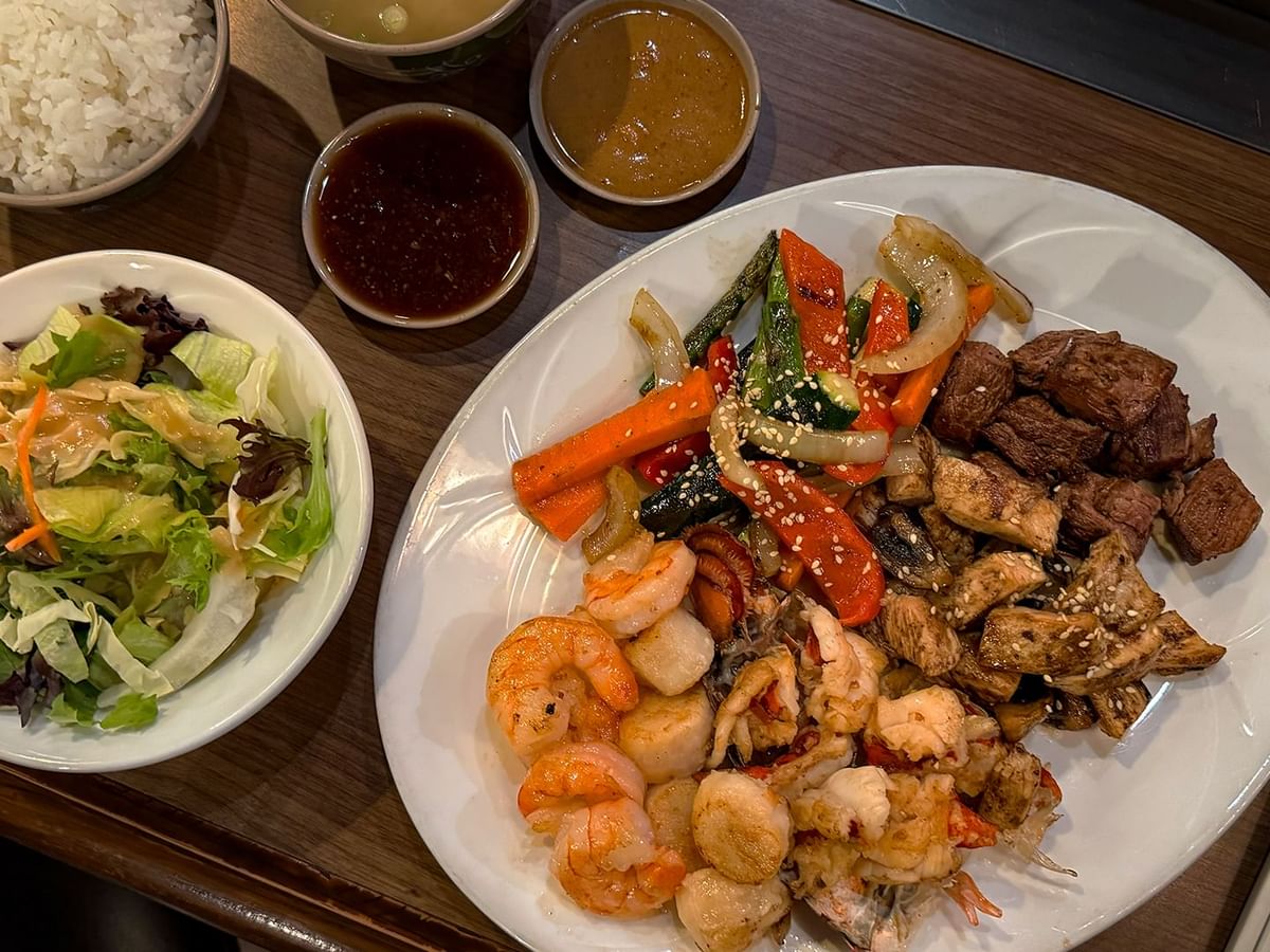 A plate of mixed seafood and meat with sauces, salad, rice, and chopsticks on a wooden table.
