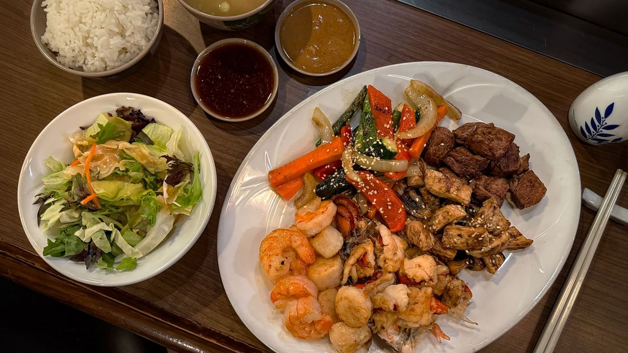 A plate of mixed seafood and meat with sauces, salad, rice, and chopsticks on a wooden table.