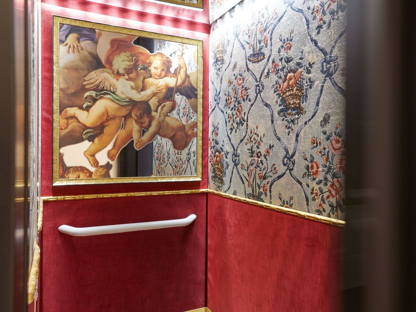Interior of an elevator with ornate wallpaper and a framed painting of cherubs.