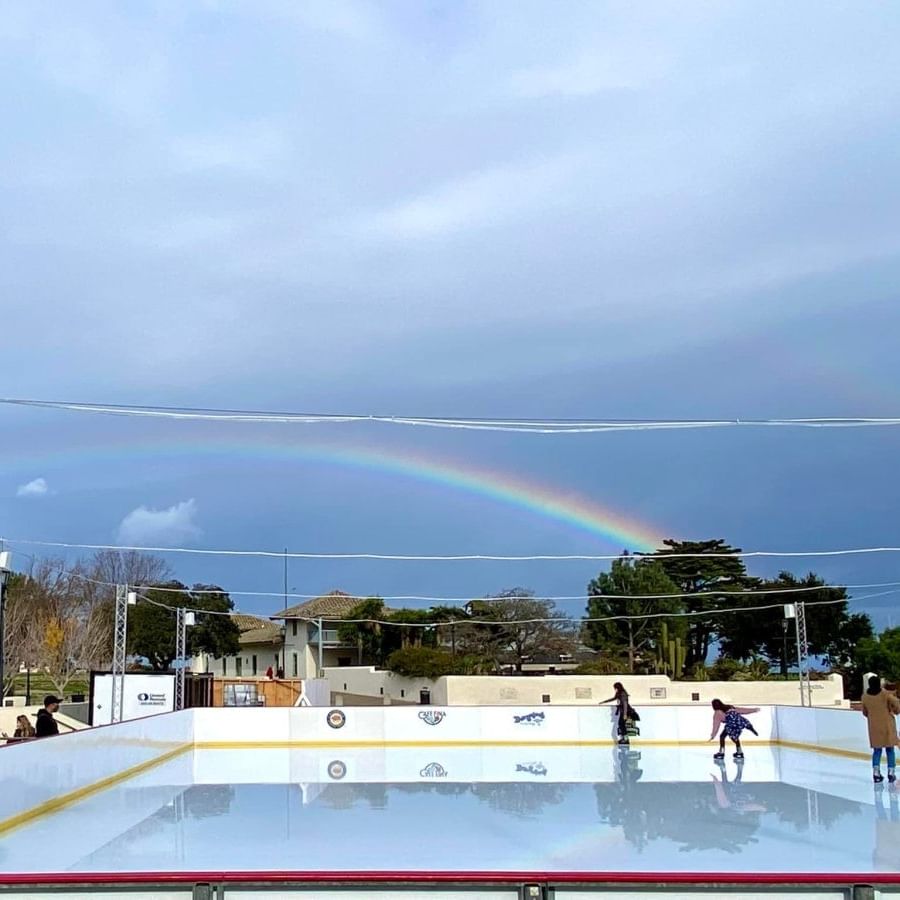 People ice skating on an outdoor rink under a rainbow with buildings and trees in the background.