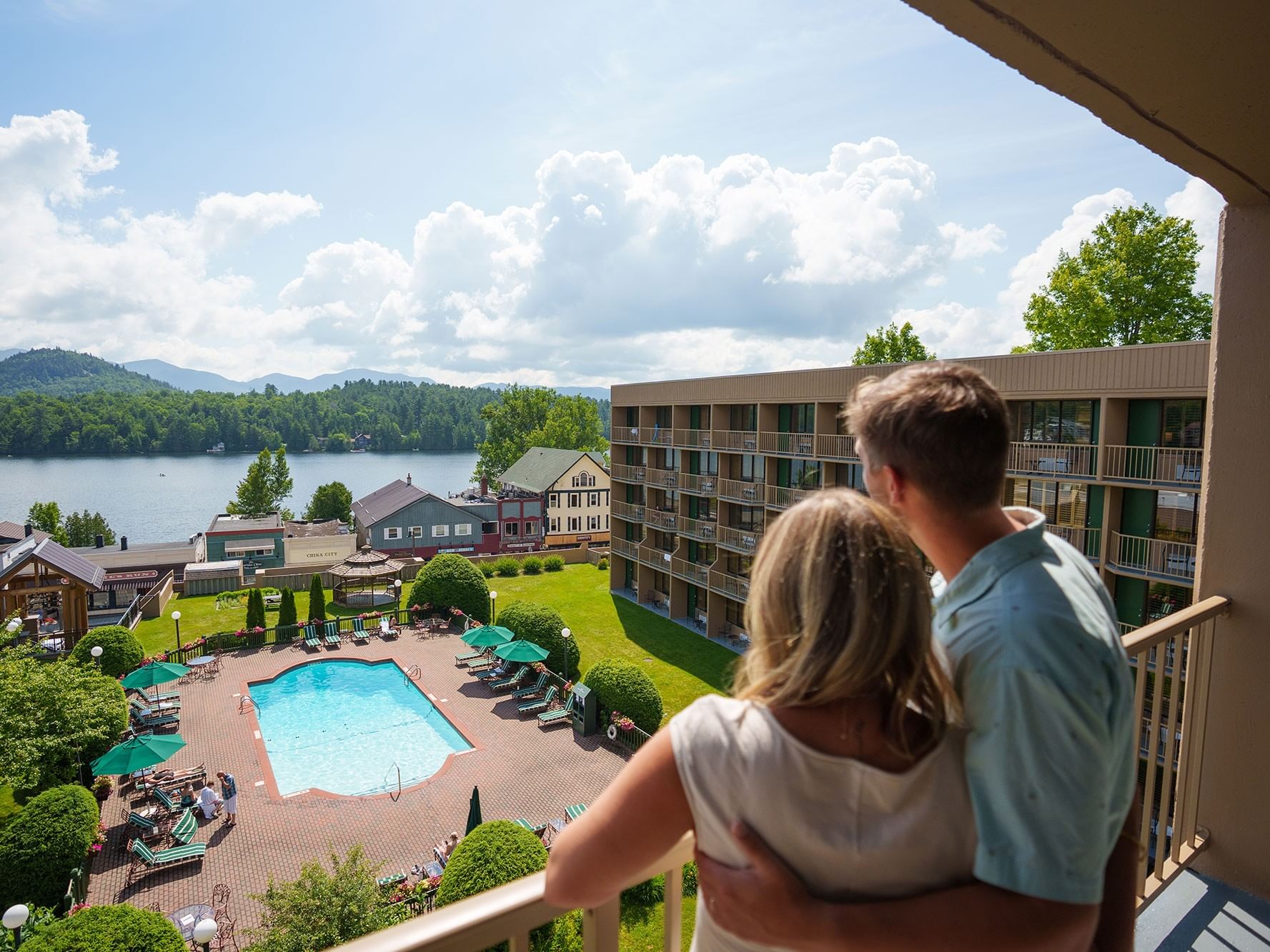 Couple on balcony overlooking outdoor pool, lake, and mountains under a bright blue sky with fluffy white clouds.