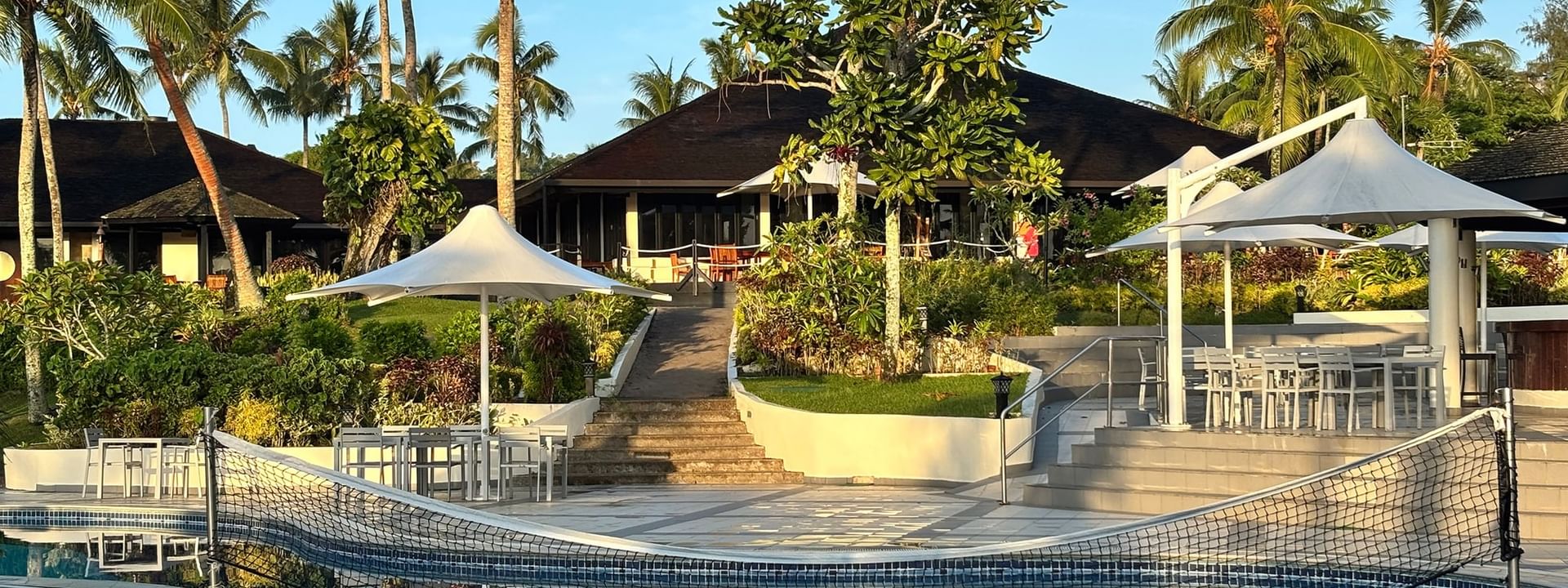 Swimming pool with a net by dining tables under white umbrellas at Warwick Le Lagon - Vanuatu in Efate