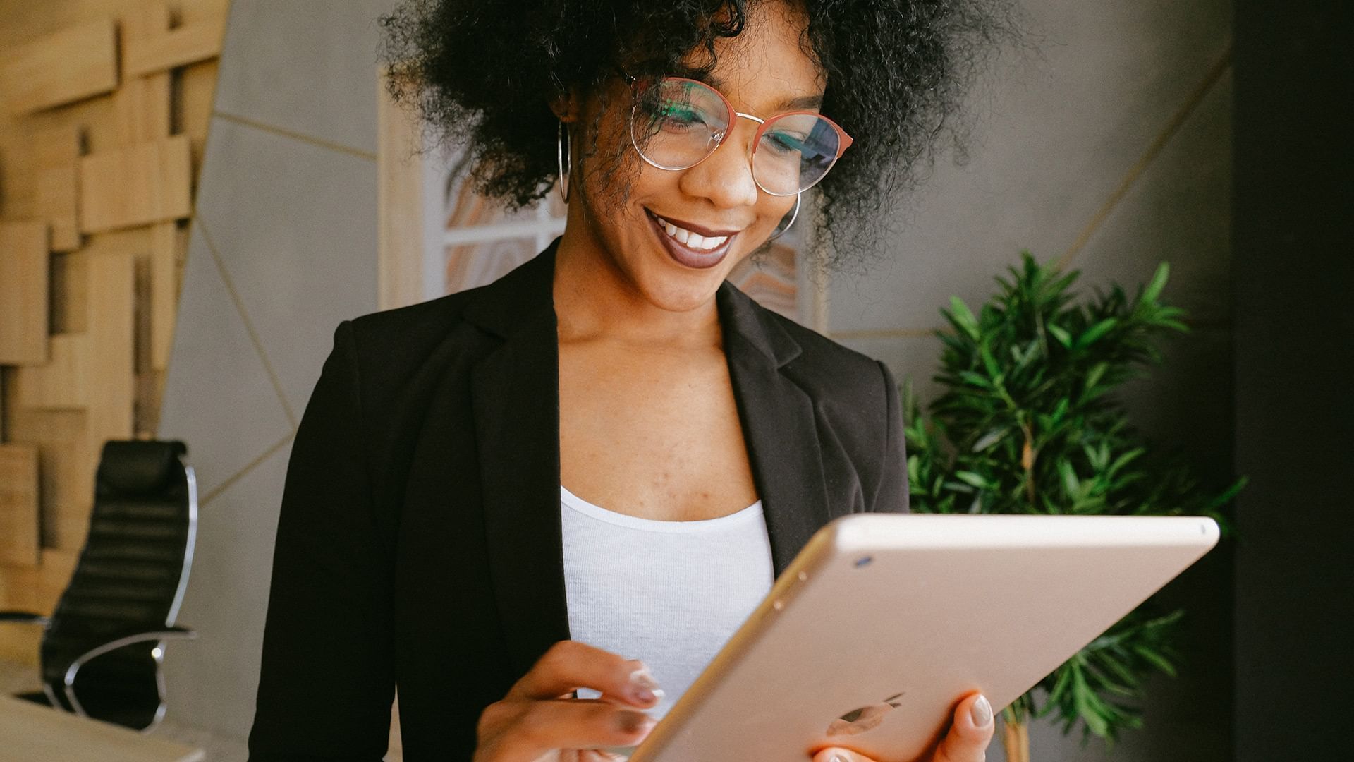Woman in blazer and glasses smiling while holding a tablet in an office.
