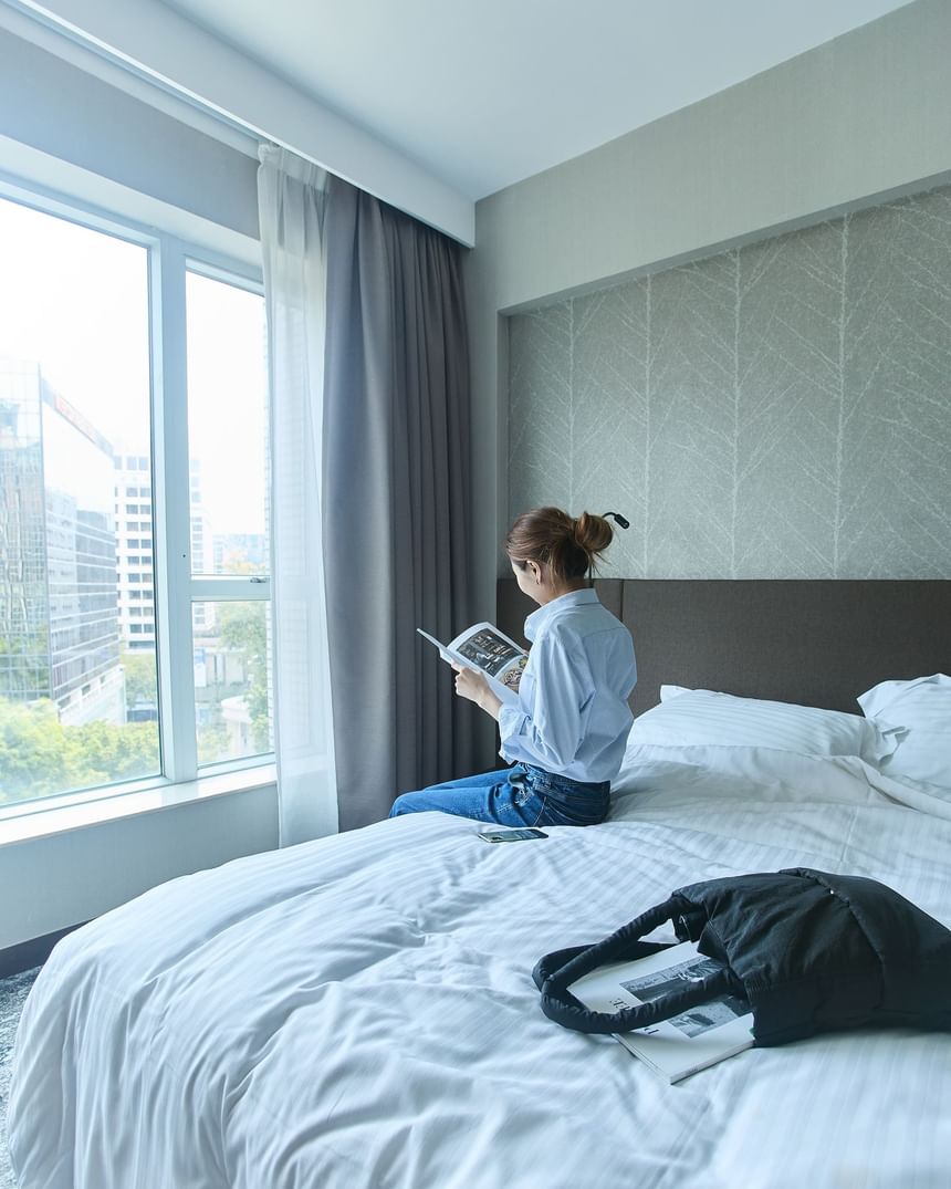 住客坐在香港百樂酒店尖沙咀高級客房的床上，在大窗旁閱讀書刊，享受城市景觀