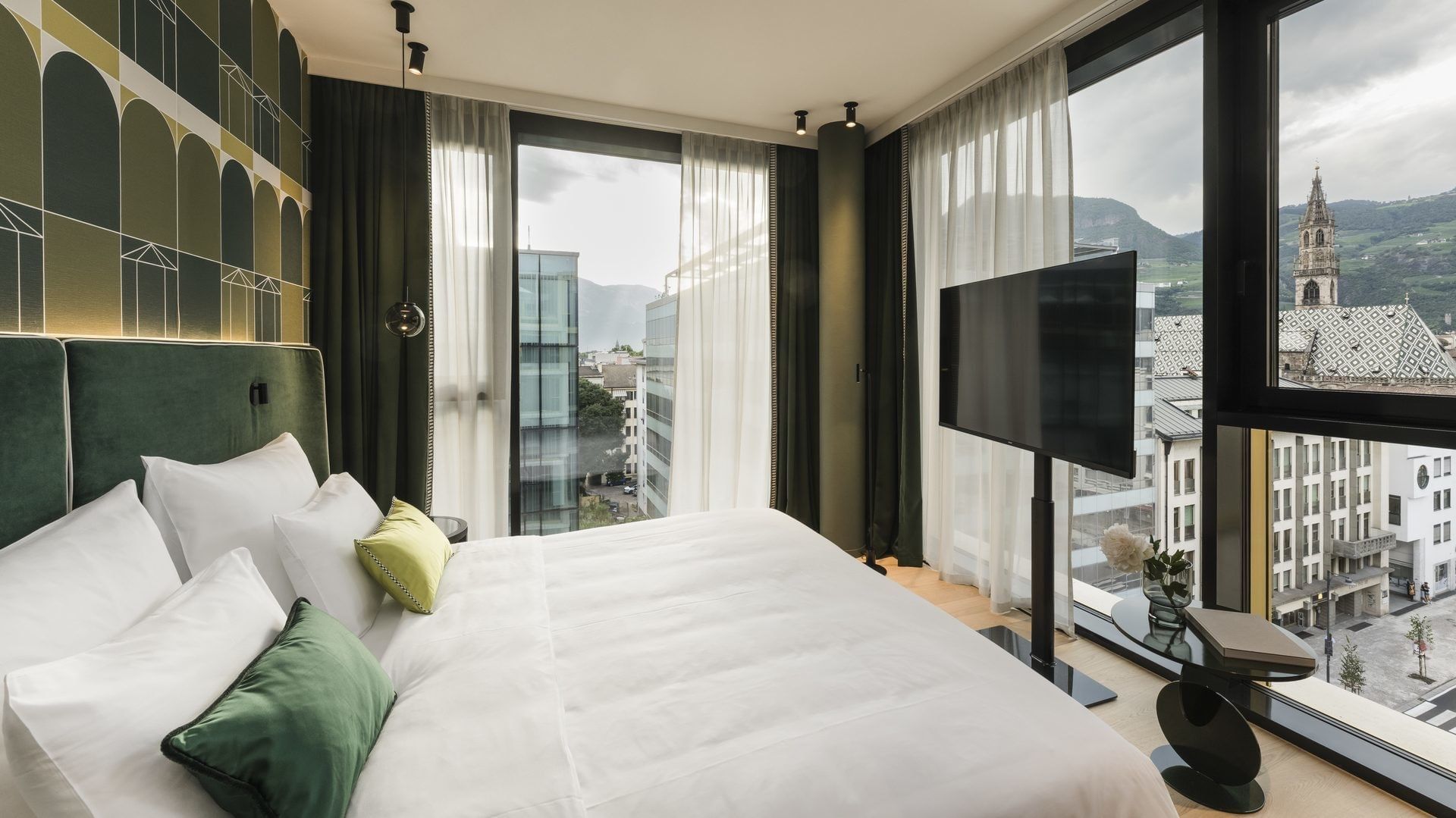 White bed with green pillows and blanket, green headboard, and city view through large windows.