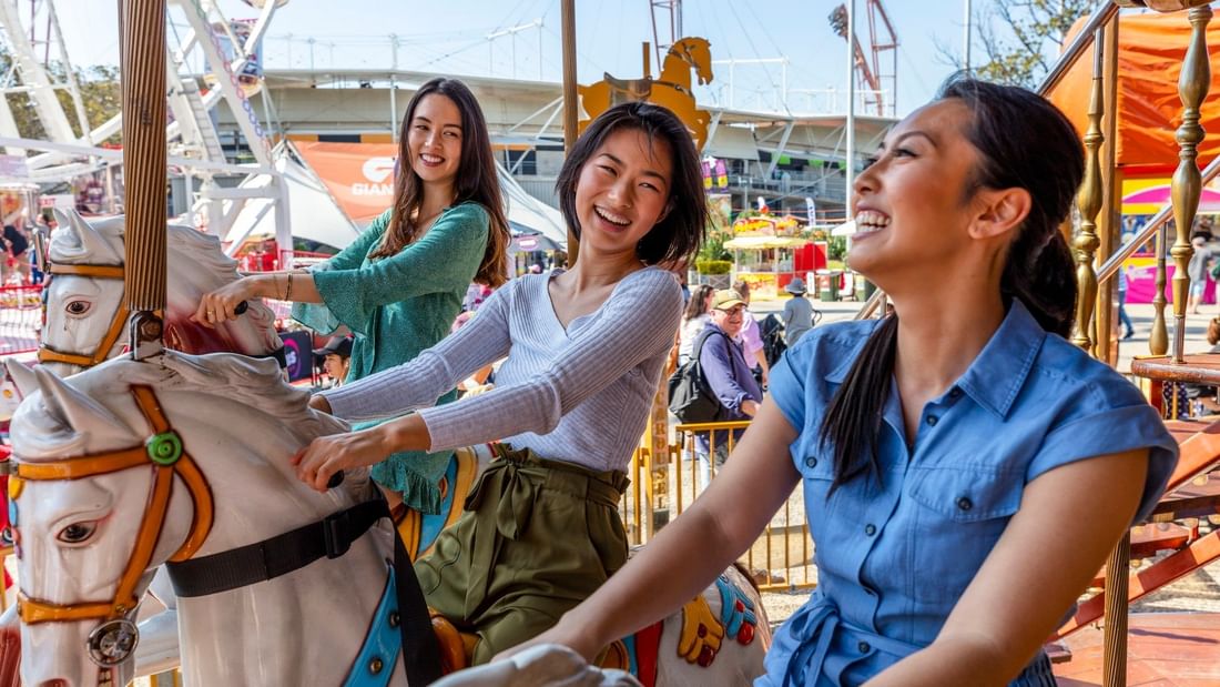 Three women smiling and riding horses on a merry-go-round at Sydney Royal Easter Show 2026.