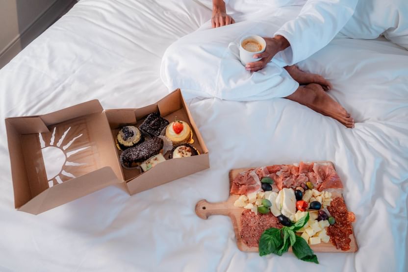 Person in white robe on bed with coffee, dessert box, and charcuterie board.