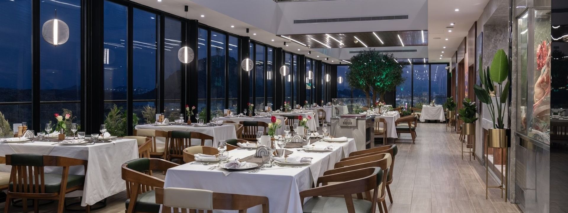 Dining tables by tall windows under a wood-slat ceiling with linear lights in Karkan Restaurant at Warwick Al Taif Hotel