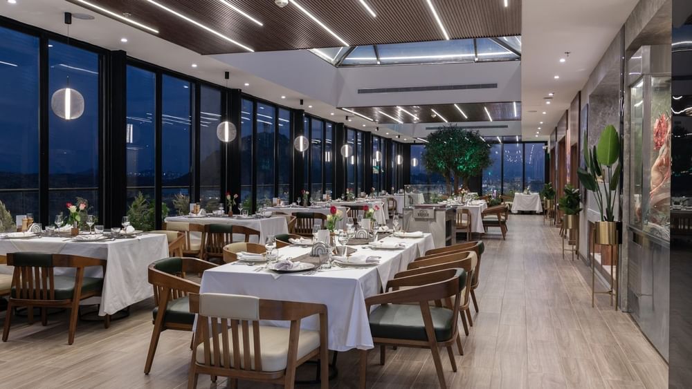 Dining tables by tall windows under a wood-slat ceiling with linear lights in Karkan Restaurant at Warwick Al Taif Hotel