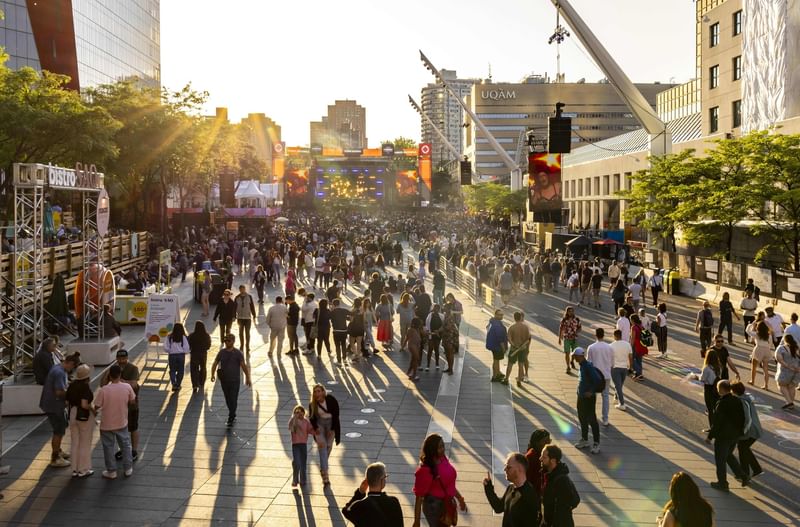 Crowds gather at an outdoor festival plaza during golden hour with stage screens and urban buildings at Warwick Le Crystal Montreal