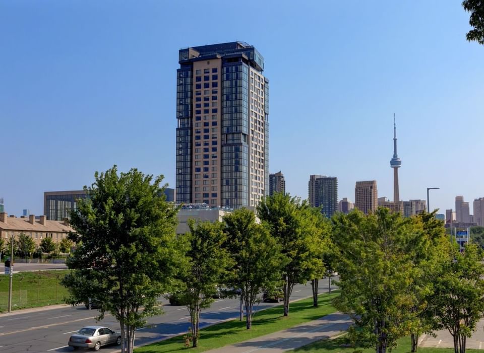 Exterior of Hotel X Toronto rising above green park trees facing distant CN Tower under clear blue sky