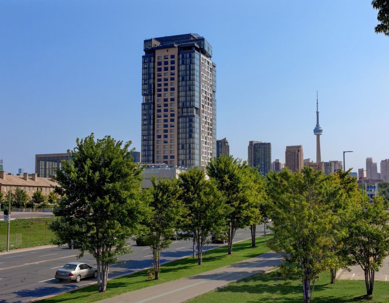 Exterior of Hotel X Toronto rising above green park trees facing distant CN Tower under clear blue sky