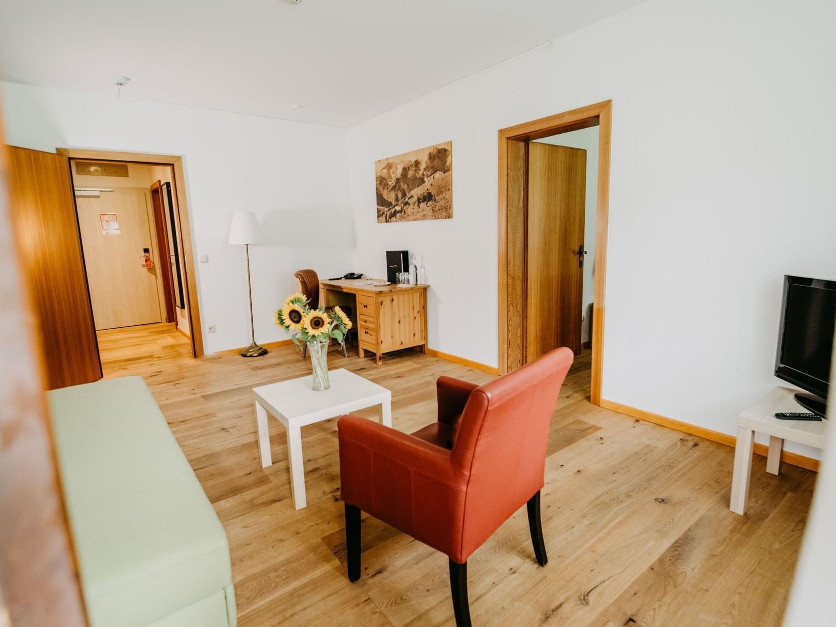 Wohnzimmer mit Holzboden, orange Sessel, Couch, Schreibtisch, Fernseher und Sonnenblumen in einer Vase.