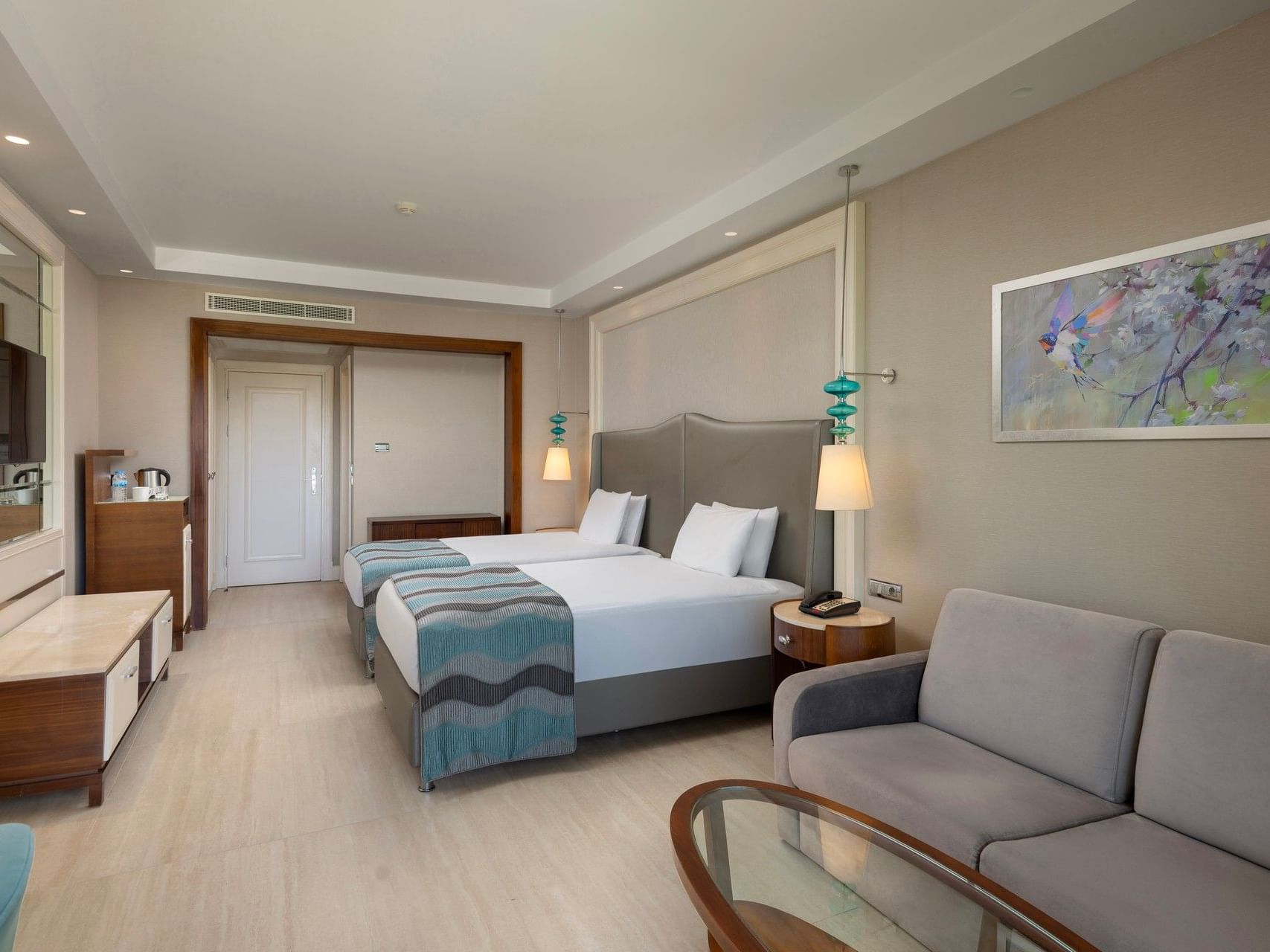 Modern hotel room with two beds, gray sofa, wooden furniture, and floral artwork at Titanic Deluxe Golf Belek