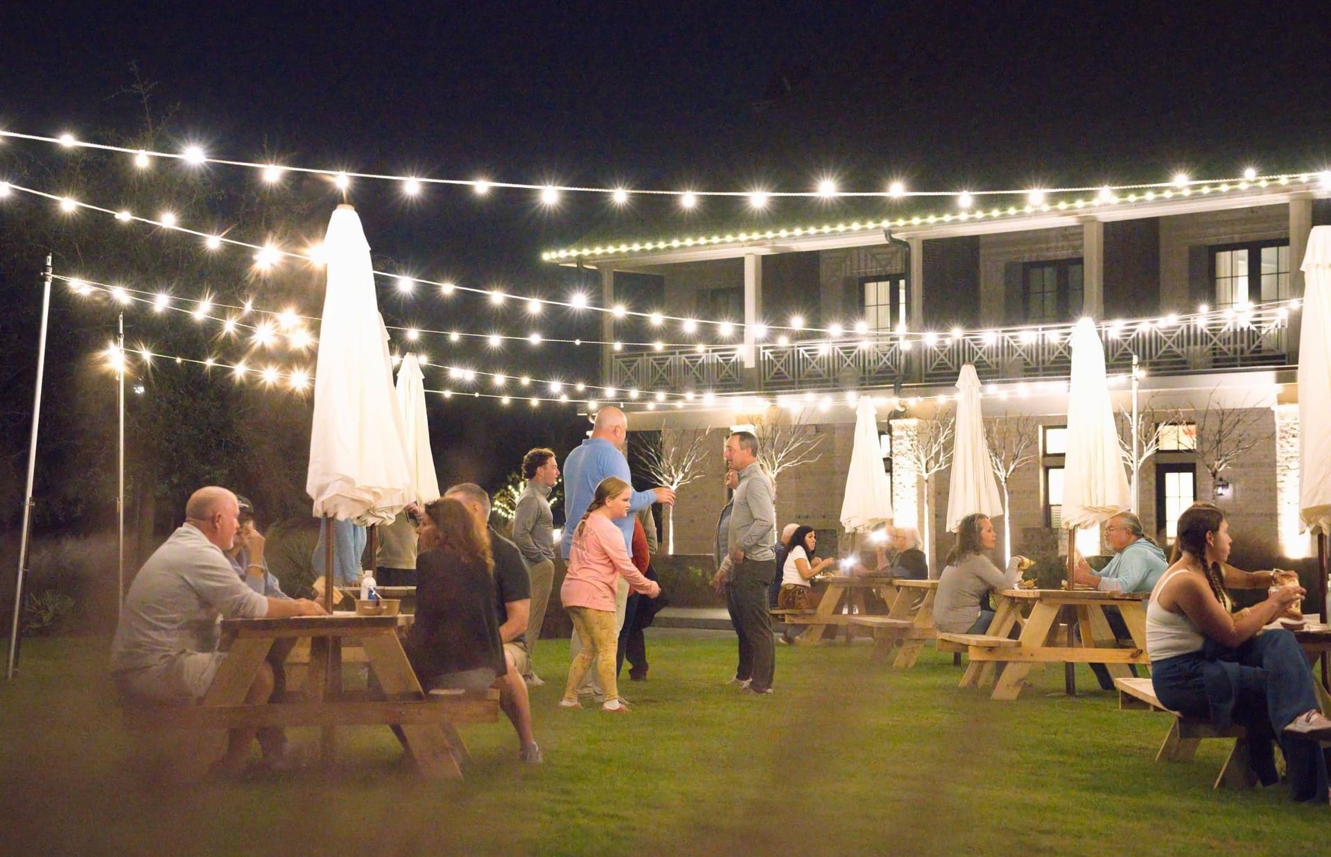 People gather at outdoor tables under string lights at night in front of a building with balconies.