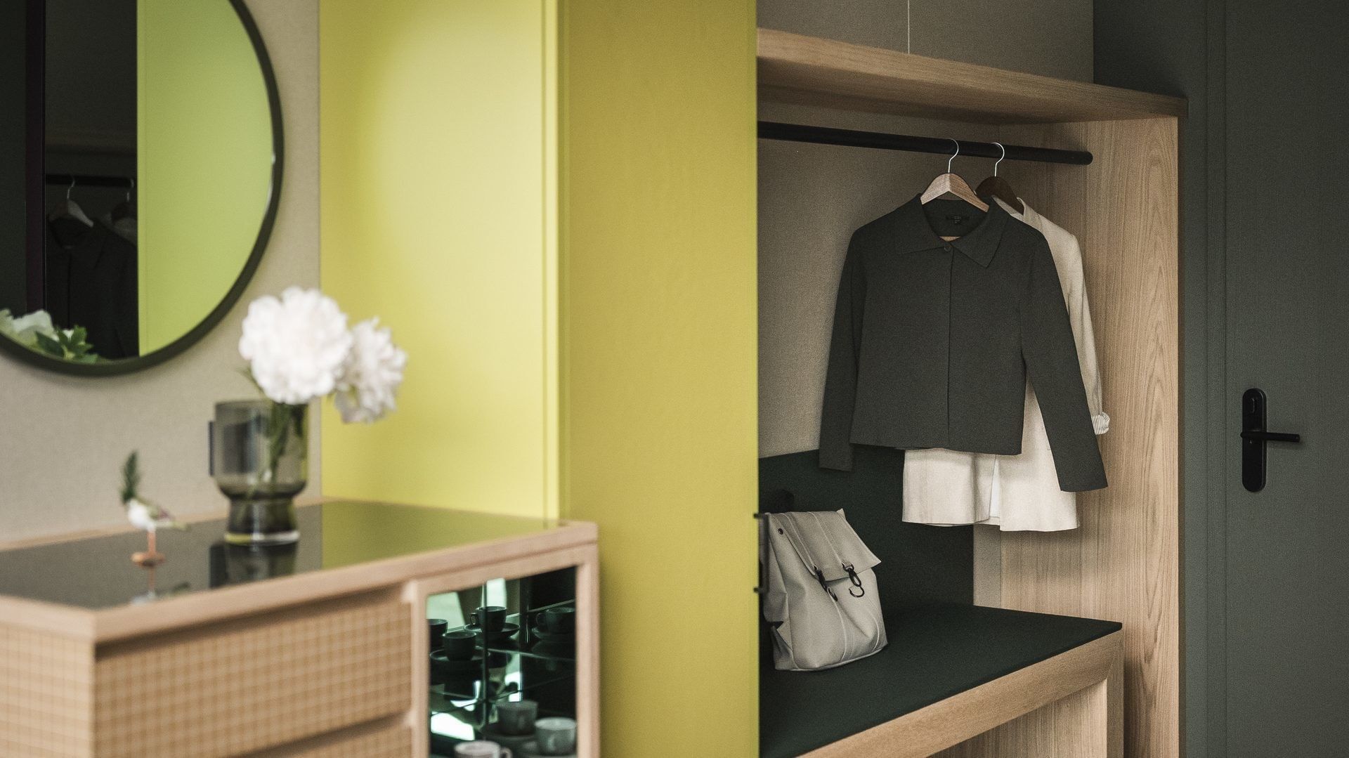 Zimmer mit Kleiderschrank, hängenden Kleidern, Bank, Kommode mit Vase und rundem Spiegel.