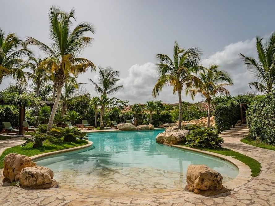 Tropical resort pool with natural rock features surrounded by palm trees and lush landscaping under a partly cloudy sky.
