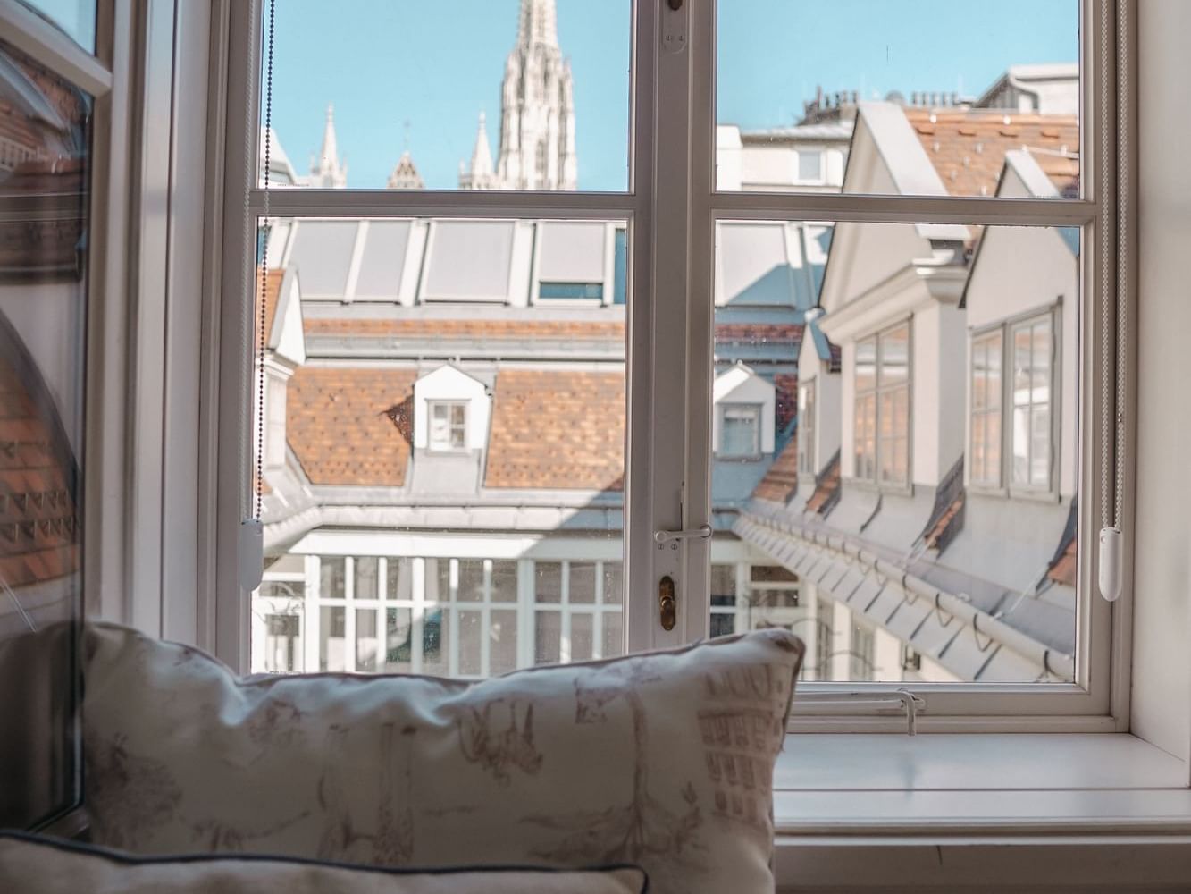 Two pillows on window sill with view of city rooftops and steeple under blue sky.
