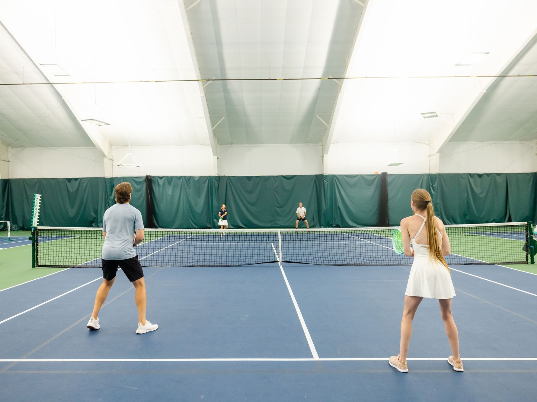 Two people playing tennis indoors with nets and courts in background.