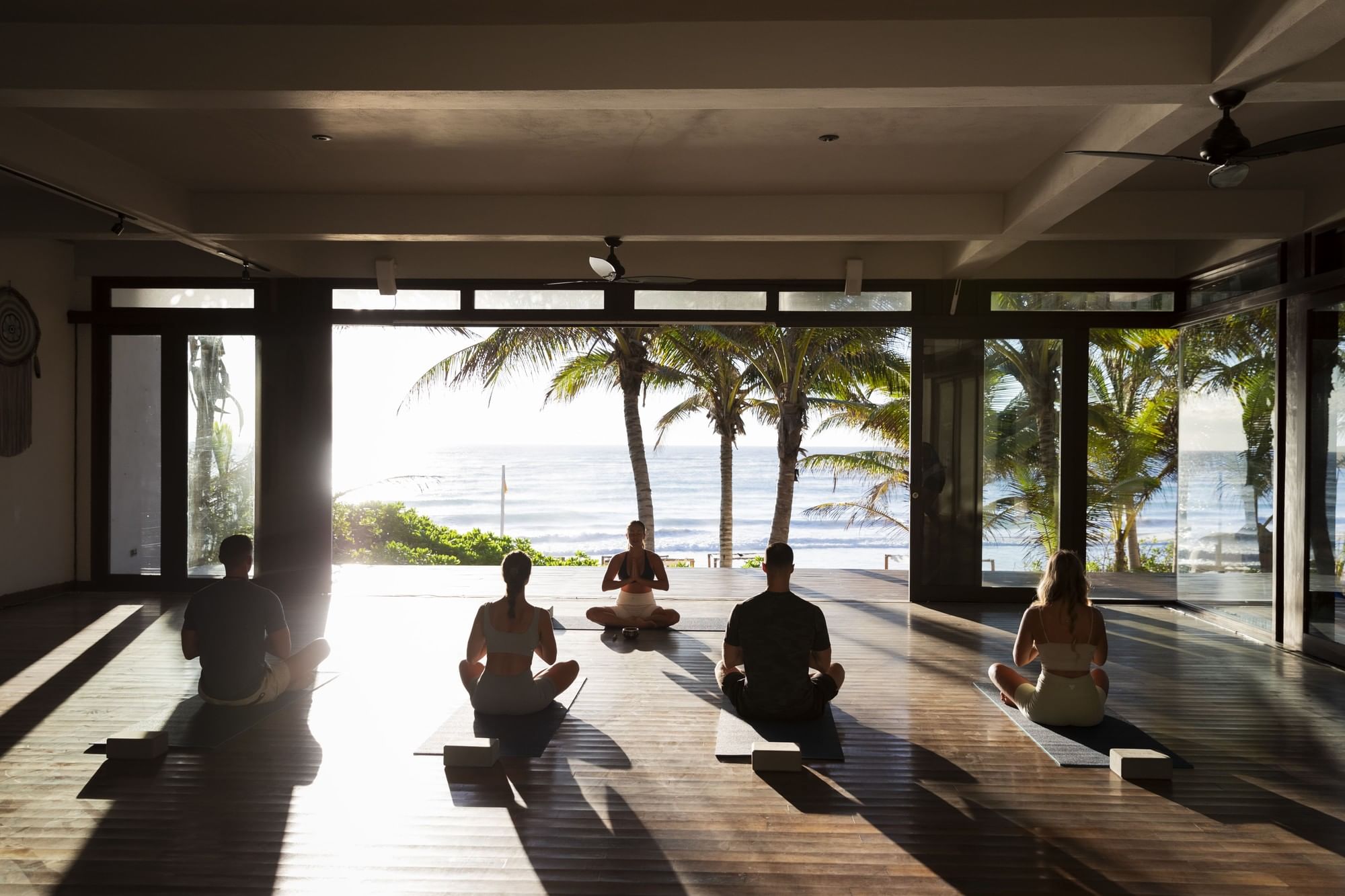Sunlight streams into the open-air yoga studio at La Zebra Hotel where five people meditate facing the ocean and palm trees