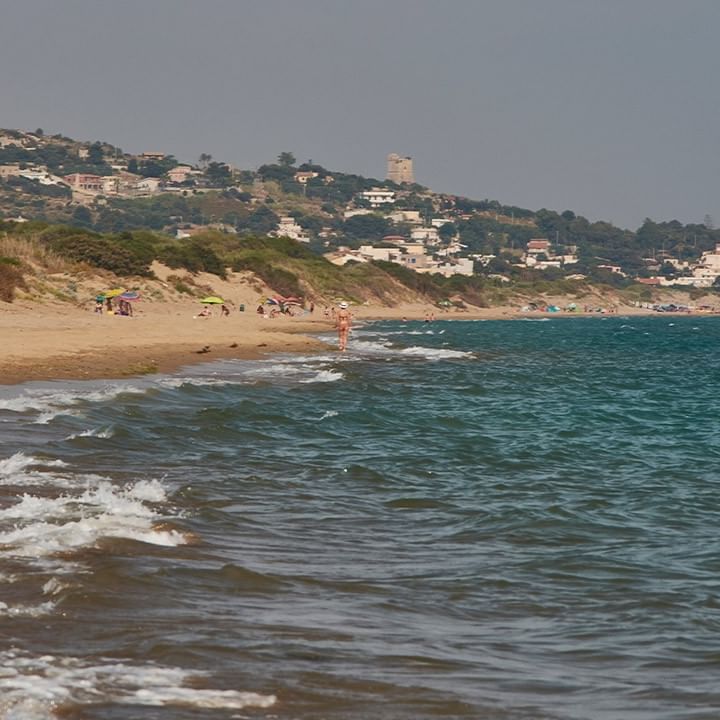 Strand mit sanften Wellen und Gebäuden am Hügel im Hintergrund, bevölkert von einigen Menschen.