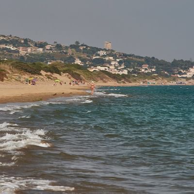 Strand mit sanften Wellen und Gebäuden am Hügel im Hintergrund, bevölkert von einigen Menschen.