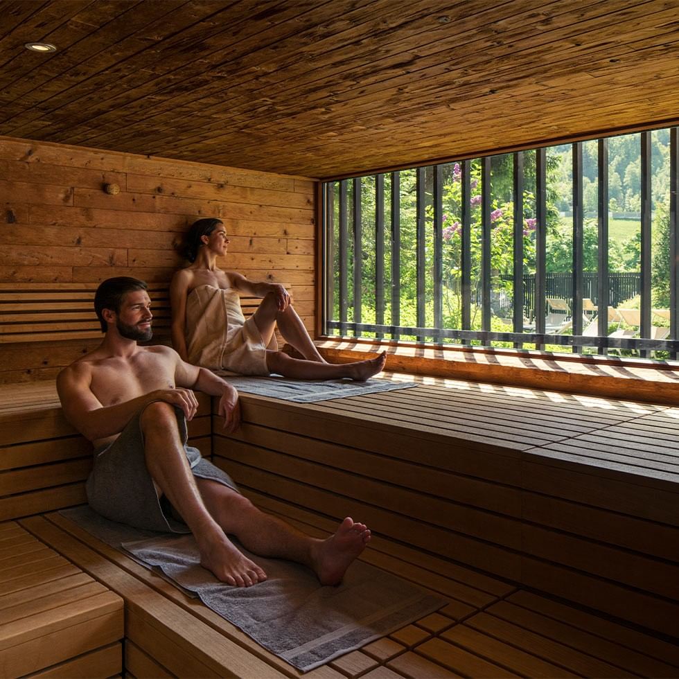 A man and a woman relax in a wooden sauna with a large window overlooking greenery.