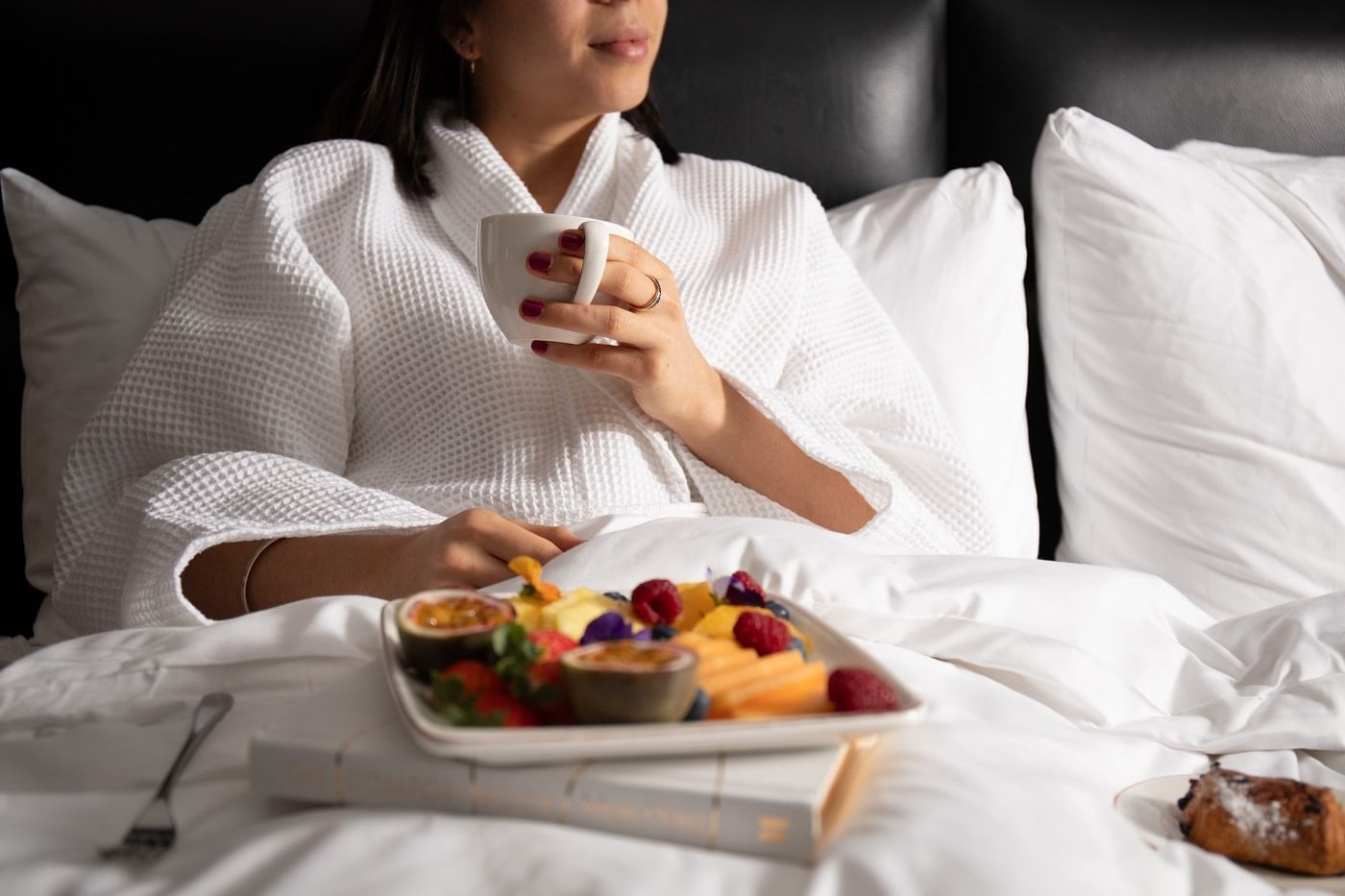 Woman in a white robe enjoying breakfast in bed with a mug and a tray of assorted fruits at The May Fair Hotel