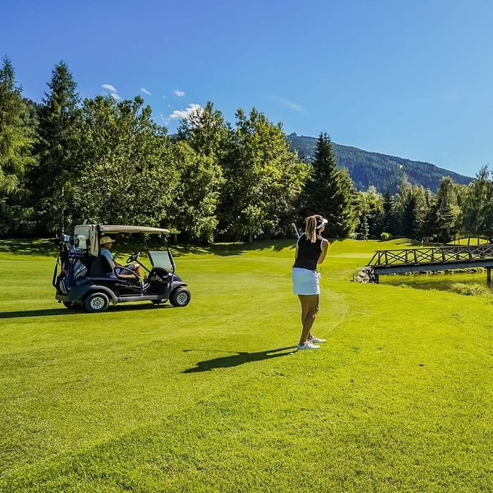Golferin steht auf Rasenplatz neben Golfcart, umgeben von Bäumen und Berg im Hintergrund.