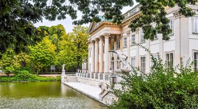 Elegant historical building with columns and statues beside a pond, surrounded by green foliage near Polonia Palace Hotel
