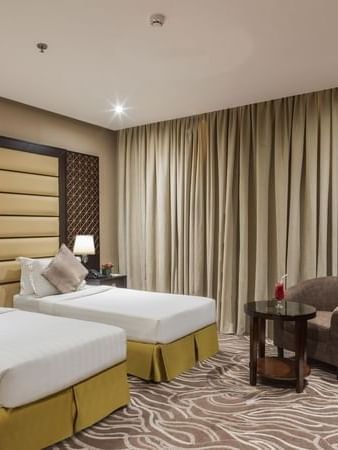 Twin beds by a round table with a drink under a long tan curtain in Deluxe King / Twin Room at Naviti by Warwick Al Khobar