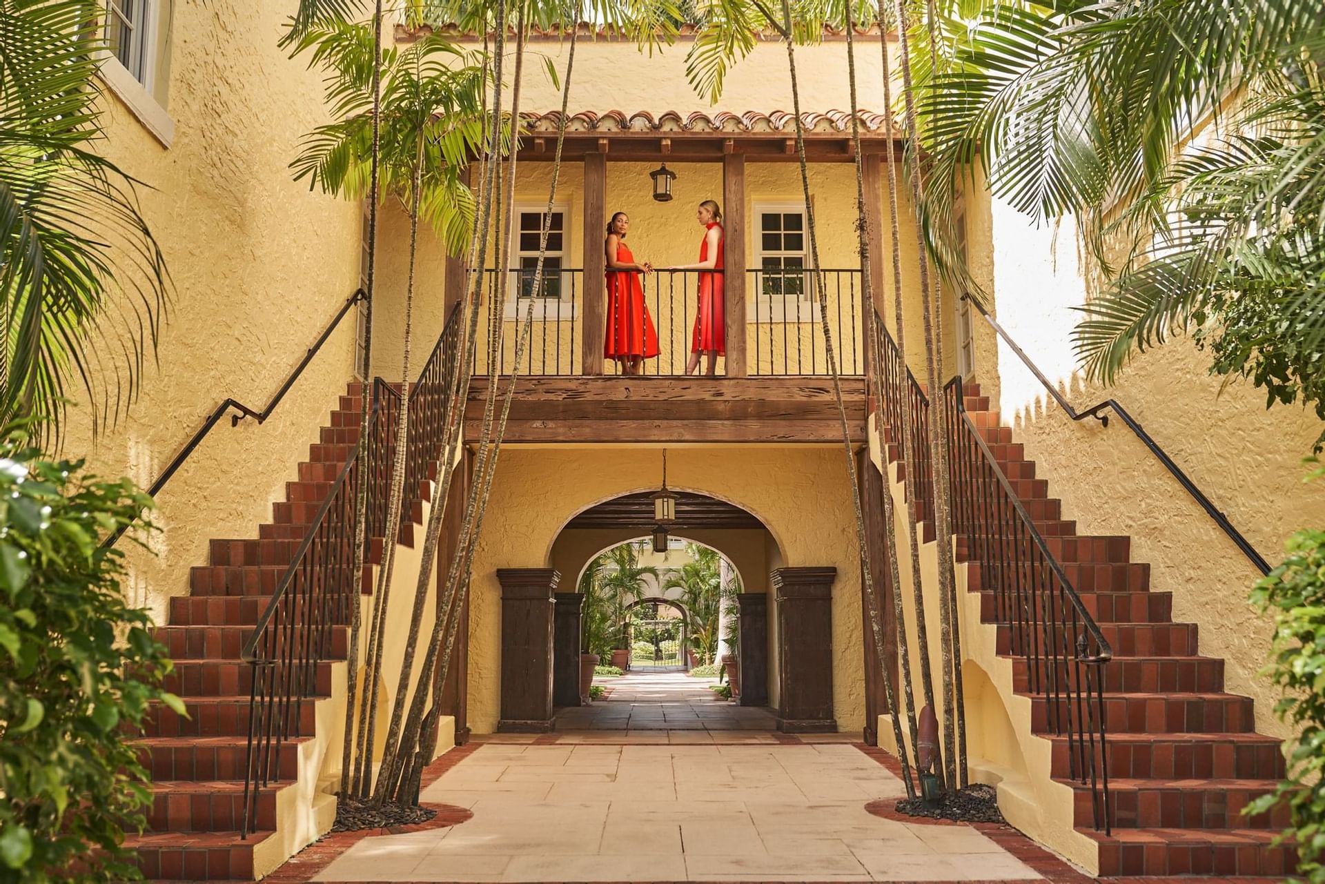 Two women in red dresses stand on a balcony above a courtyard with stairs and palm trees.