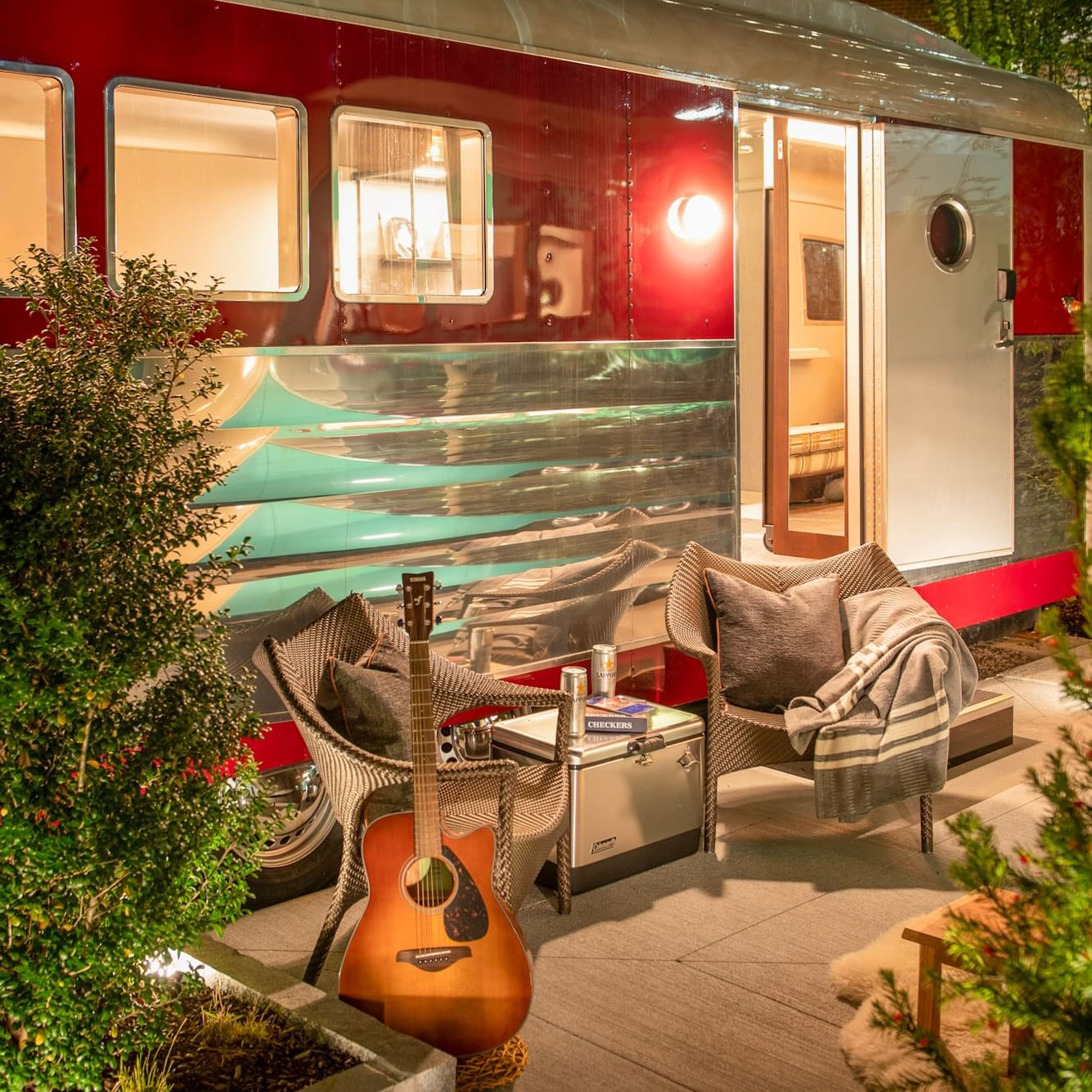 Illuminated vintage trailer with red and turquoise exterior, outdoor seating area, guitar, and surrounding greenery at dusk.